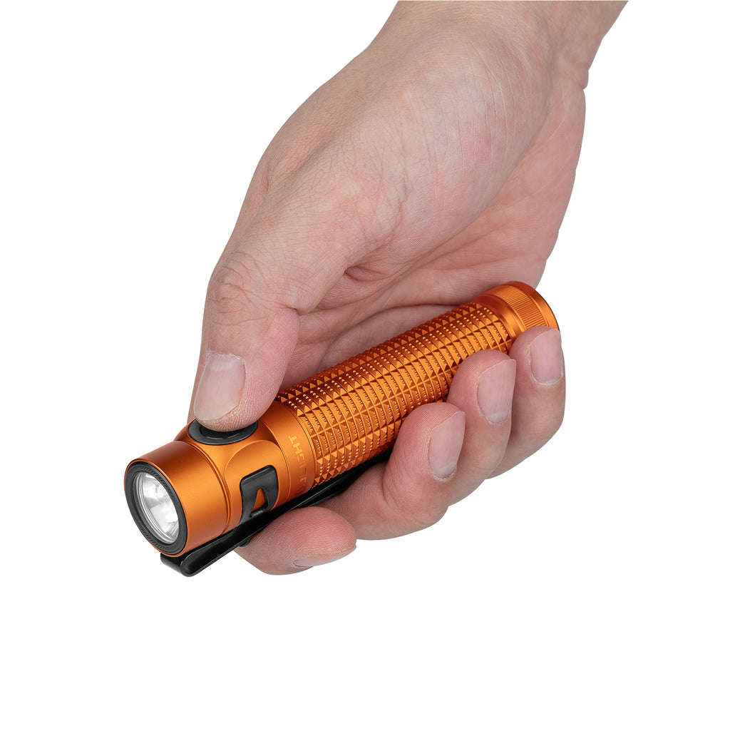 Hand holding an orange flashlight with a textured grip, black clip, and side button against a white background.